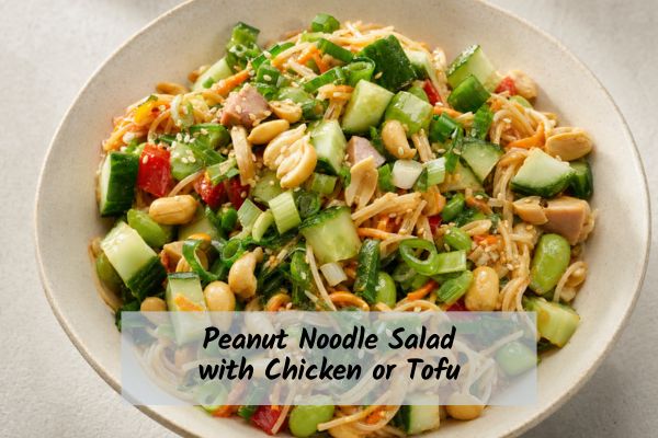 Peanut-Noodle-Salad-Chicken-Tofu-Recipe A vibrant bowl of peanut noodle salad topped with chunky cucumber pieces, whole roasted peanuts, green onions, edamame, diced red peppers, shredded carrots, and thin noodles, garnished with sesame seeds and labeled "Peanut Noodle Salad with Chicken or Tofu."