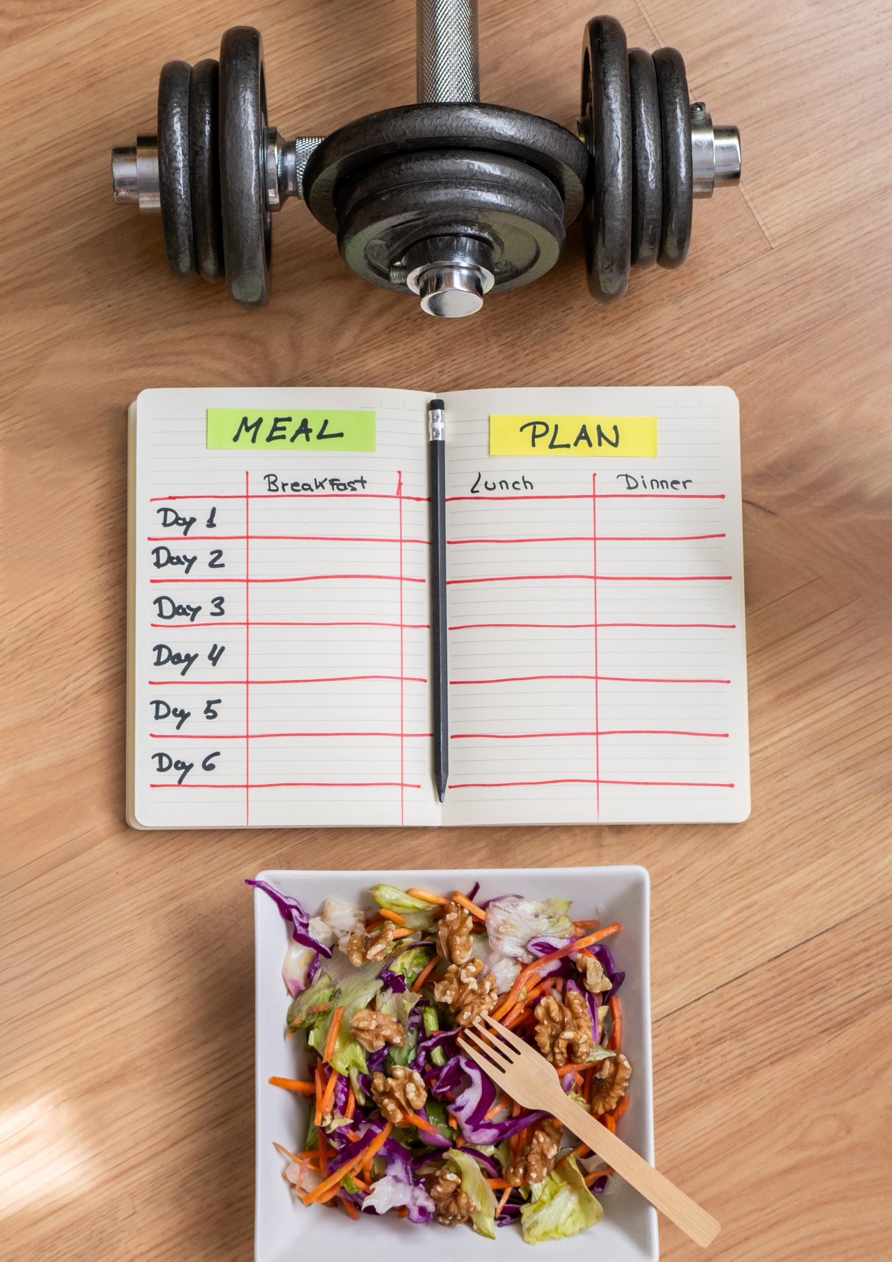 Fitness enthusiast is preparing a healthy meal plan, accompanied by dumbbells and a nutritious salad, symbolizing the combination of exercise and balanced nutrition for a wellness lifestyle a meal plan journal in front of weights with food on the ground